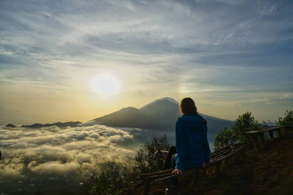 Martina von Travelgrapher sitzt bei Sonnenaufgang am Rand des Kraters des Mount Batur und beobachtet die Sonne hinter dem Gunung Abnag und dem Batur Lake aufgehen